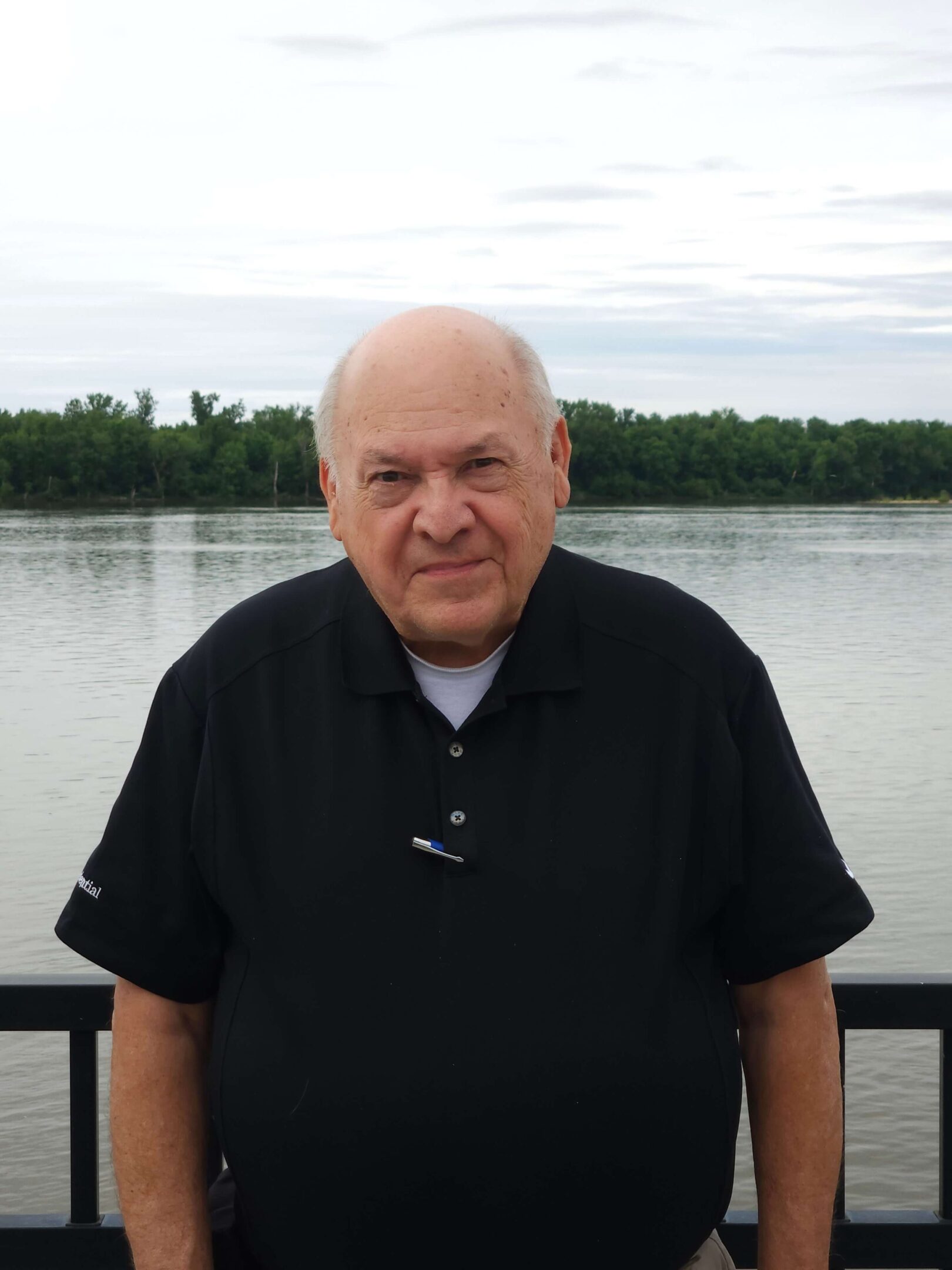 Elderly man standing by a lakeside railing.